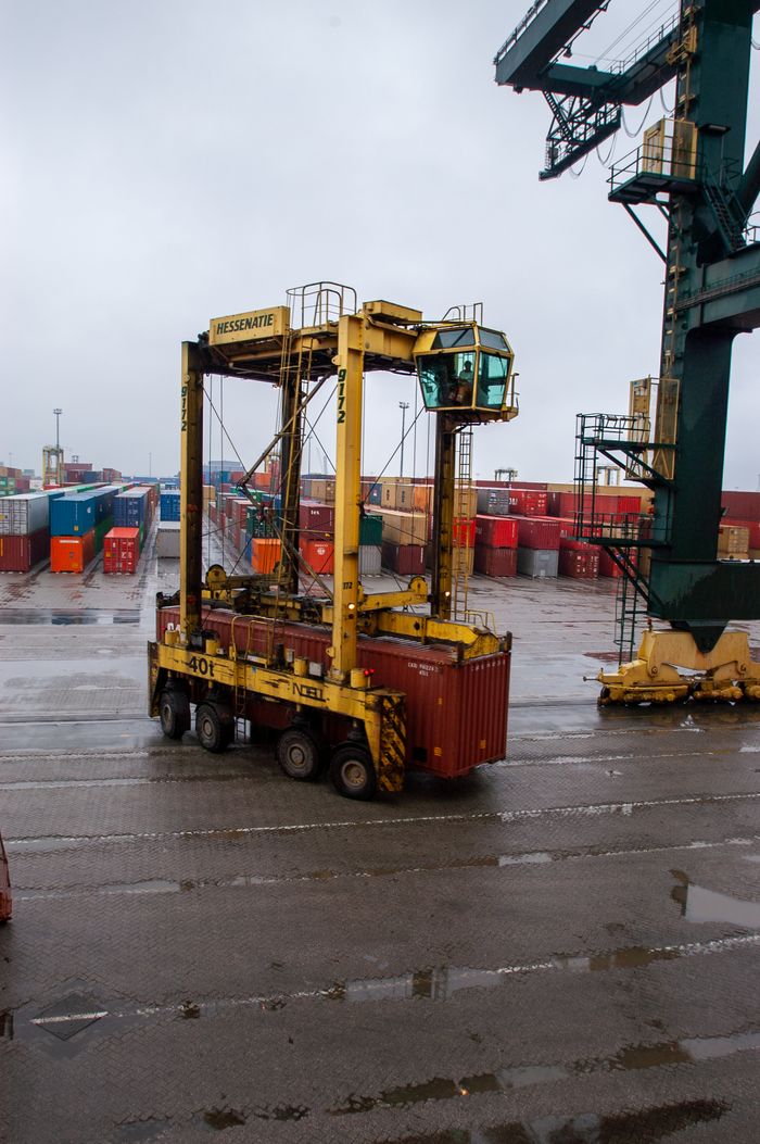 Yellow straddle carrier moving containers at a busy, rainy shipping port.