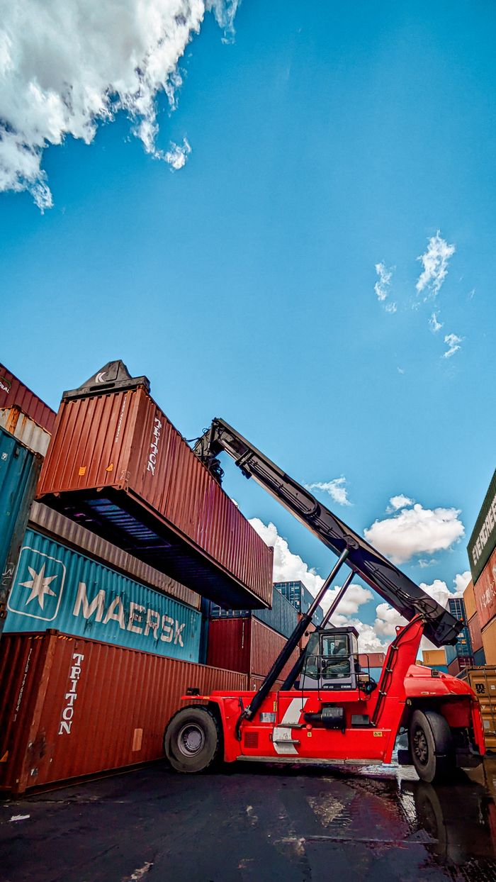 Red hydraulic crane handling shipping containers at a port under a clear blue sky.