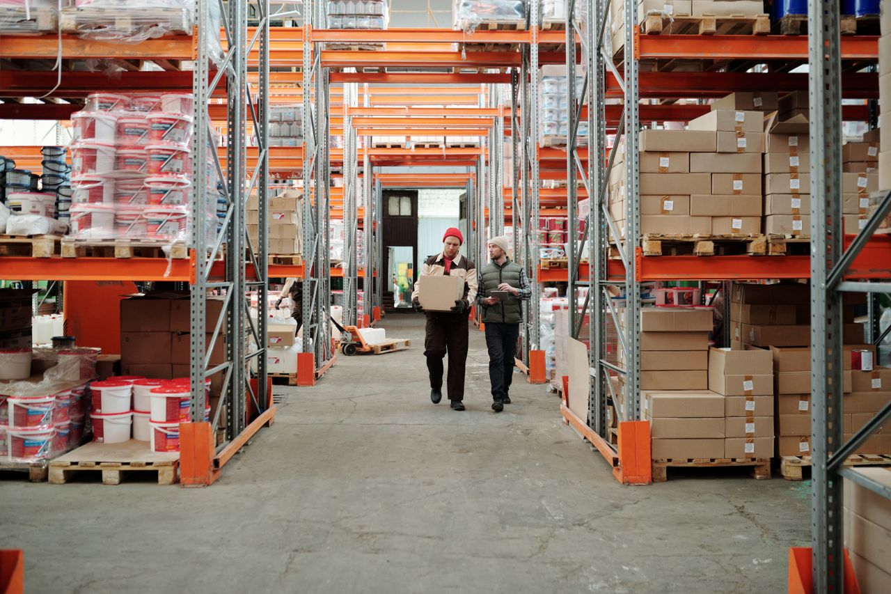 Home Two workers handle a package in a spacious warehouse surrounded by shelves stocked with boxes and products.